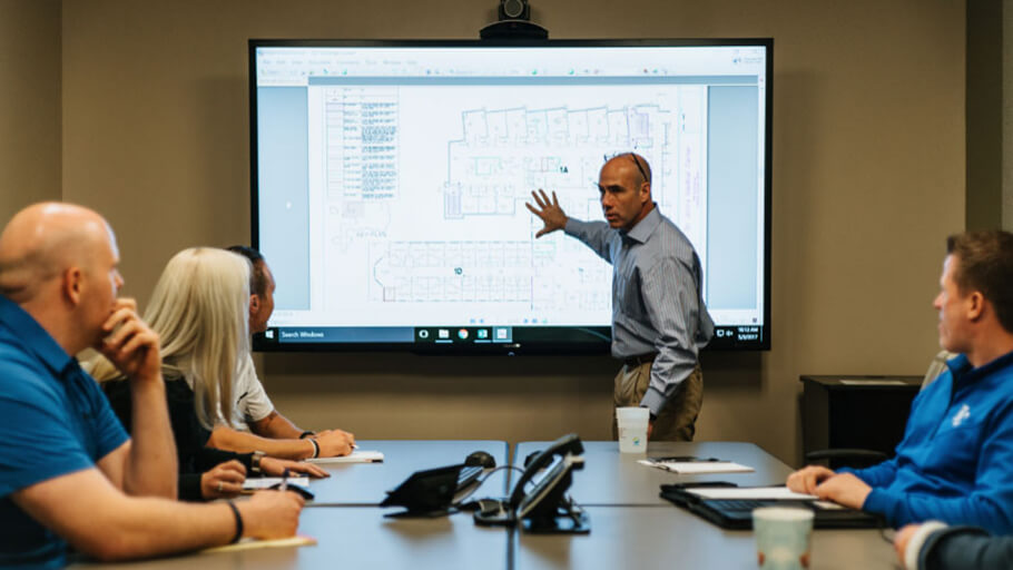 Darrell Stein presenting Henderson Engineers service offerings on a large TV to a room full of engineers in a conference room.